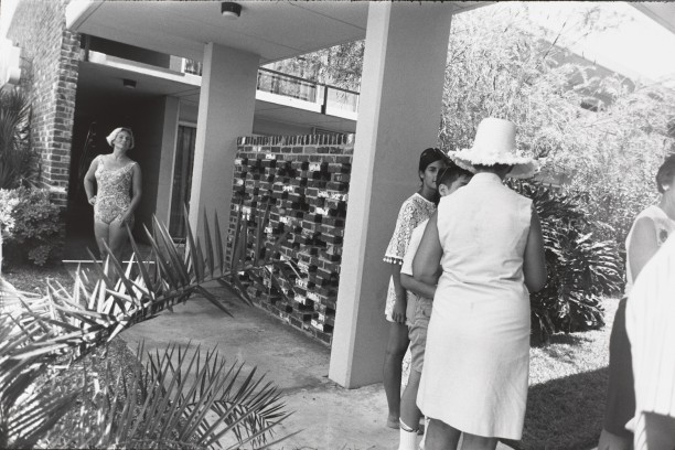 Untitled (Woman in bathing suit standing at building entrance, in foreground is a group with backs to camera)